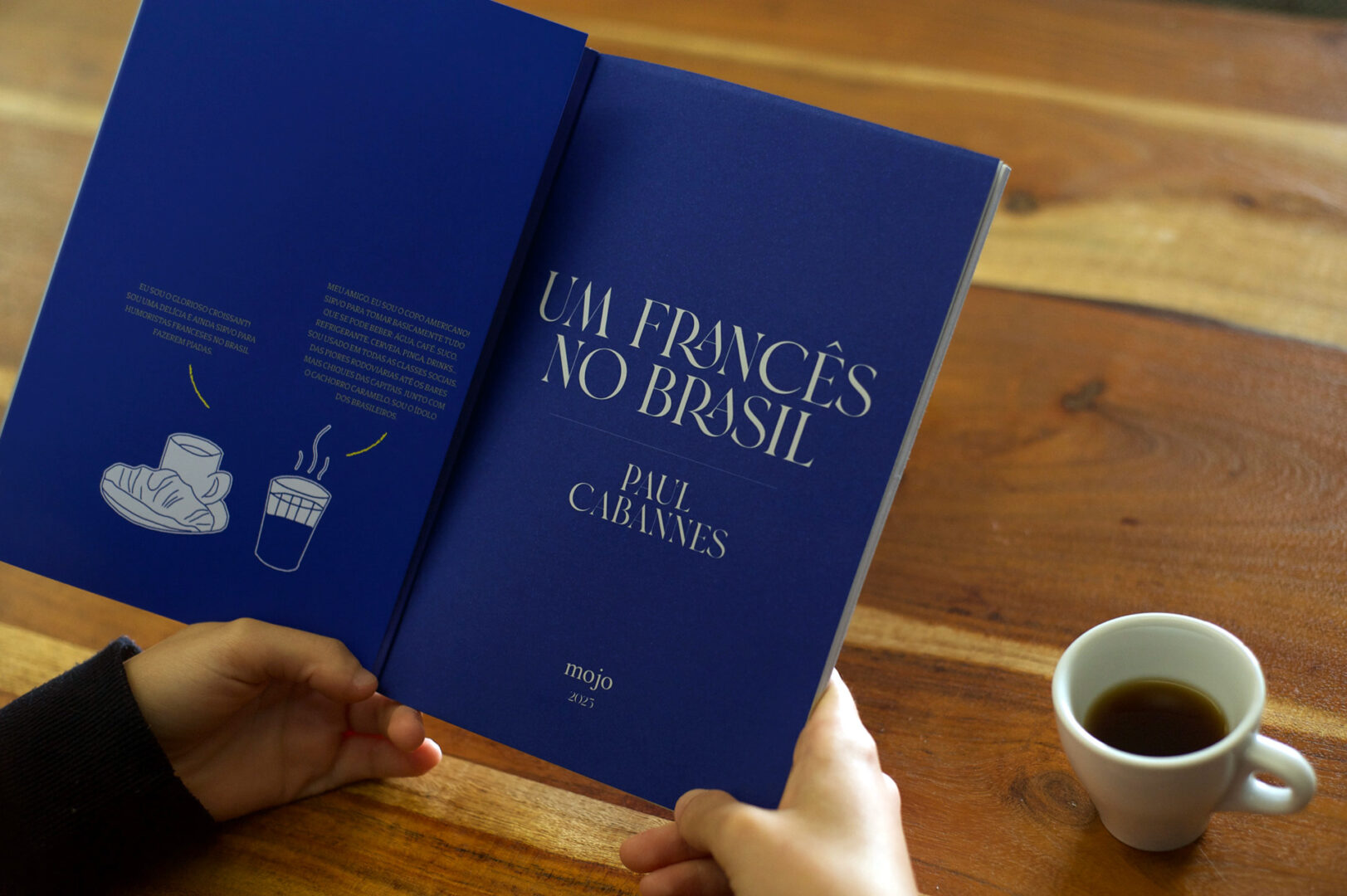 Livro aberto sobre a mesa, com o título em branco sobre azul e as badanas desenhadas a traço